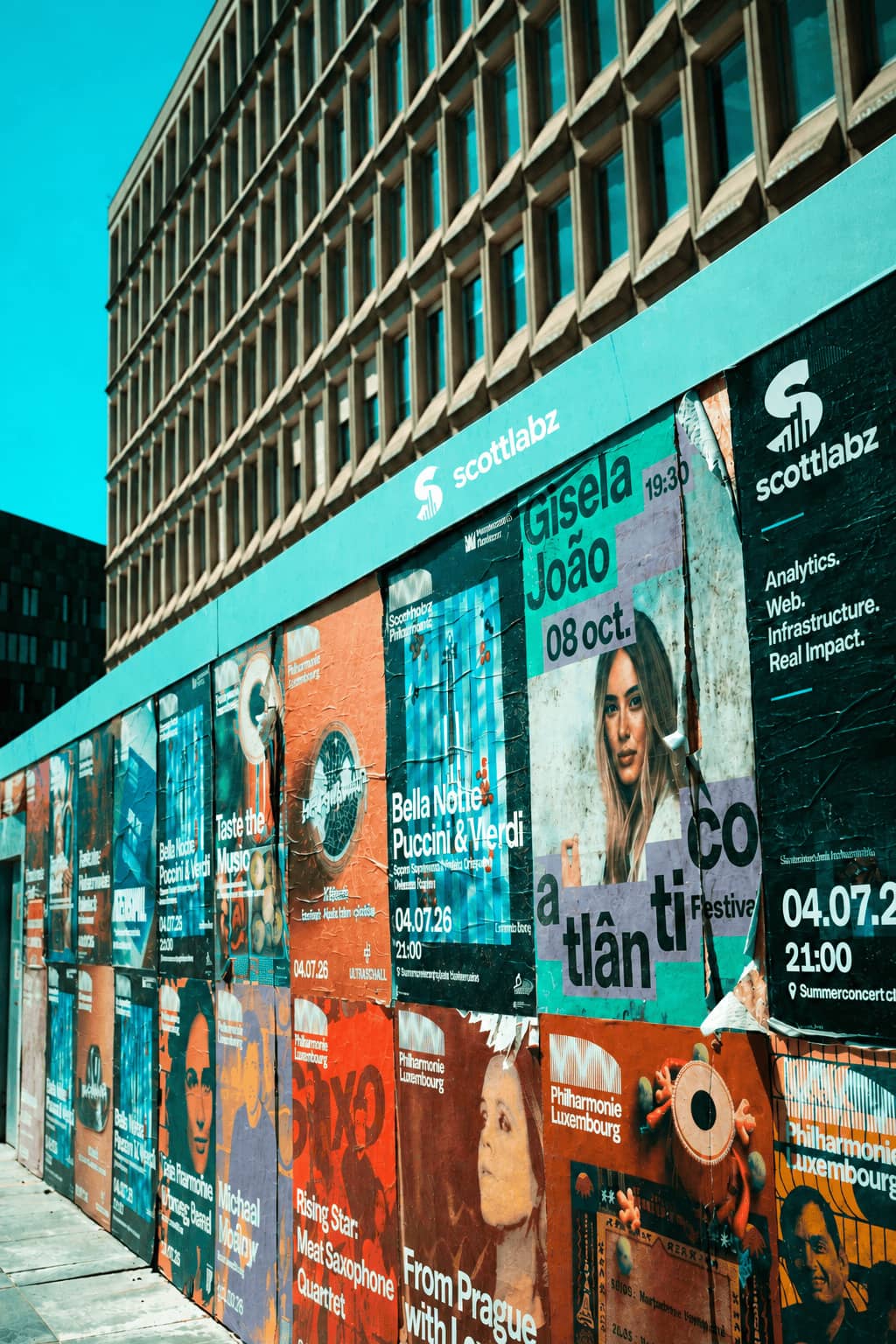 Street-level wall of layered concert and event posters featuring a Scott Labz ad reading 'Analytics. Web. Infrastructure. Real Impact.', representing advertising and media buying services